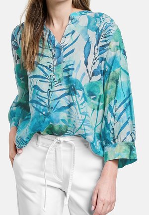 Frau trägt eine locker sitzende, langärmelige Bluse mit blau-grünem botanischem Muster, die in weiße, mit Kordelzug versehene Hosen gesteckt ist.