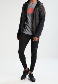 Idrottskläder som inkluderar en grå t-shirt med röd bergslogga, svart zip-up jacka, svarta leggings och svarta löparskor.