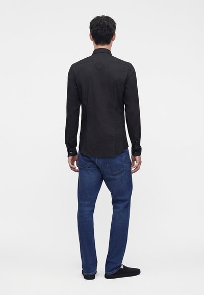 Mann trägt ein schwarzes Langarmshirt, blaue Jeans und schwarze Schuhe und steht mit dem Rücken zu einem schlichten weißen Hintergrund.