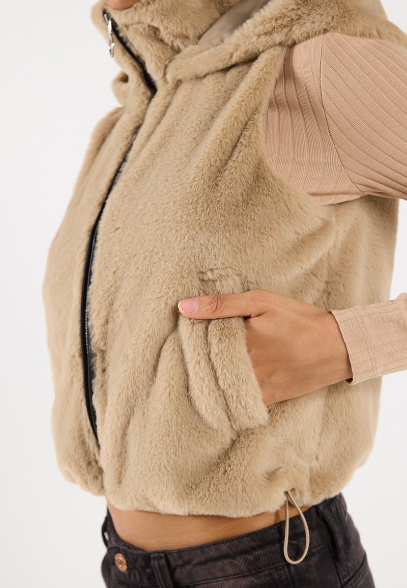 Beige nepbont gilet met een capuchon, rits aan de voorkant en zijzakken. Het heeft een zachte textuur en een verstelbare trekkoord aan de onderzijde.