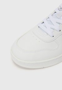 Sneaker bianca con tomaia in pelle liscia, punta tondeggiante e lacci in tessuto. Suola testurizzata con scanalature sottili e perforazioni vicino alla punta.