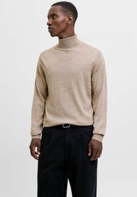 Beżowy sweter w golf z miękkiego materiału dzianinowego, z długimi r�ękawami i ściągaczami, zestawiony z ciemnymi spodniami. Prosty, neutralny design.
