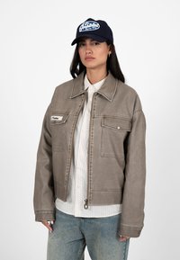 Kvinde iført en grå jakke med lynlås over en hvid skjorte med striber, lyseblå jeans og en marineblå kasket med hvid tekst.