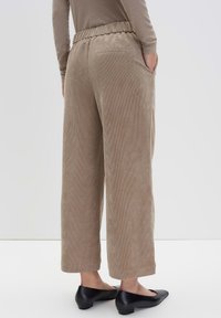Personne portant un pantalon en velours côtelé beige à jambes larges et coupe cropped, avec une taille élastique et des chaussures à petit talon noires, se tenant avec les mains dans les poches.