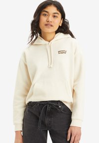 Ung kvinna iklädd en off-white hoodie från Levi's med framficka och dragsnöre, tillsammans med höga svarta jeans som är knutna i midjan.