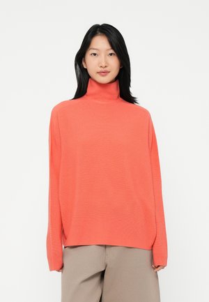 Donna con capelli neri lisci che indossa un maglione a collo alto corallo largo e pantaloni beige a gamba larga in piedi contro uno sfondo bianco.