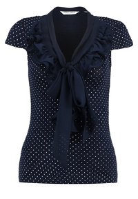 Navyblå blus med kort ärm, vita prickar, volangkrage och en knuten rosett framtill, gjord av mjukt tyg.