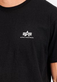 Camiseta de algodón negra con cuello redondo, mangas cortas y un logotipo impreso en blanco con un estilo de letra estilizada en la parte superior izquierda.