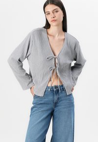 Gray textured top with long sleeves and a plunging V-neck, featuring a tie closure at the front and a relaxed fit. Paired with blue jeans.