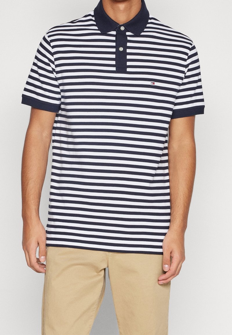 Homme portant un polo à rayures horizontales bleu marine et blanches avec un col bleu marine et un pantalon beige, debout devant un fond blanc.