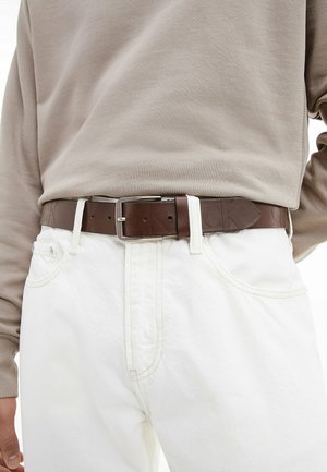 Środkowa część ciała osoby ubranej w beżową, długorękawową koszulę wpuszczoną w białe jeansy zapięte paskiem z brązowej skóry.