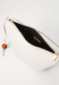 Hellbeige Lederhandtasche mit goldenem Reißverschluss, beigem Innenfutter und einer dekorativen bernsteinfarbenen Perle am Reißverschluss. Sie hat ein minimalistisches Design.