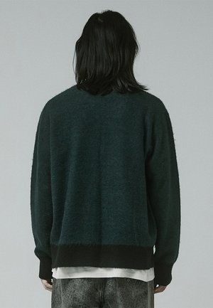 Pull oversized vert foncé avec une finition texturée. Poignets et ourlet côtelés, épaules tombantes et coupe décontractée. Porté sur une chemise blanche.