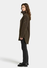 Brun fleece jakke med høj krave, ribbede mansjetter, sideslids i kanten og en struktureret lapp på venstre ærme, parret med sorte jeans og støvler.