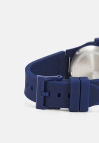 Correa de reloj de silicona azul marino con superficie texturizada, hebilla ajustable y fondo de acero inoxidable. Presenta la marca 'Hugo' en la correa.