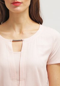 Frau trägt ein hellrosa kurzärmliges Oberteil mit einem silbernen Stangendetail am Halsausschnitt, das den Hals- und oberen Brustbereich zeigt.