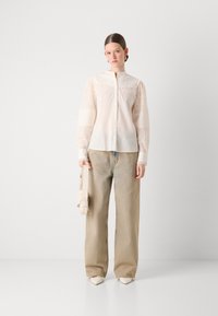 Blusa bianca di cotone con dettagli a occhio di pernice e maniche a righe, abbinata a jeans beige larghi. Il modello tiene una borsa di colore chiaro.