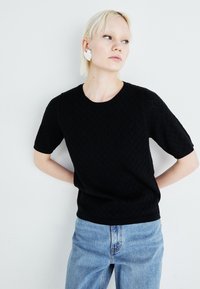 Fekete rövid ujjú pulóver, rombusz kötésmintával és kerek nyakkivágással. Világoskék, laza szabású farmernadrággal párosítva, amely egy hétköznapi öltözetet mutat be.