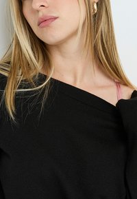 Maglione nero lavorato a maglia con design off-shoulder. Tessuto testurizzato, con una spallina di reggiseno rosa visibile sulla spalla destra. Orlo a coste sottile.