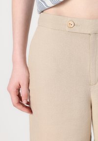 Gros plan d'une personne portant un pantalon beige taille haute avec un bouton fermé à la taille et une partie d'une chemise rayée visible au-dessus.