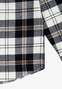 Tissu à carreaux noir, blanc et marron sur une chemise à manches longues avec un détail d’ourlet incurvé.