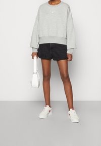 Szary sweatshirt z okrągłym dekoltem i białym logo, zestawiony z czarnymi szortami. Biała torba i białe sneakersy z czerwonym akcentem dopełniają stylizację.