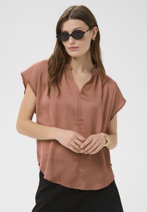 Mujer con gafas de sol ovaladas negras, blusa suelta de manga corta color óxido, falda negra y joyas doradas, de pie frente a un fondo blanco.