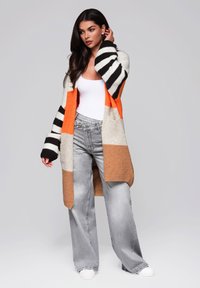 Sweter uczyński z pomarańczowymi, czarnymi i kremowymi paskami, o długim, luźnym kroju. W połączeniu z szarymi dżinsami z wysokim stanem.