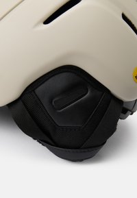 Helm mit einem matten crèmefarbenen Äußeren, schwarzer Polsterung und verstellbaren Riemen, ausgestattet mit Belüftungsöffnungen und einem gelben Logodetail an der Seite.