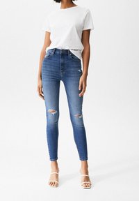 High-waisted blue denim jeans with distressed knee details and a skinny fit. Paired with a plain white t-shirt and strappy sandals.