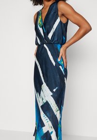 Námořnická modrá bezramínková šaty s abstraktními bílými a zelenými vzory, s designem do zavinovacího stylu a bočním rozparkem, vyrobené z hladké látky.