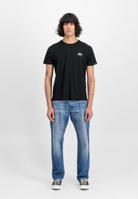 T-shirt preta de mangas curtas, decote redondo e logótipo branco no peito, combinada com jeans de perna reta de cor azul clara e ténis pretos.