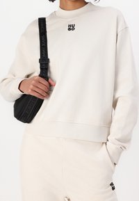 Sudadera beige con cuello y puños acanalados, que presenta un logo negro de "HUGO". Combinada con pantalones beige y un pequeño bolso cruzado negro.