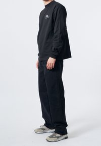 Černé tričko s dlouhým rukávem a kulatým výstřihem, zdobené bílými textovými grafikami. Spárováno s volnými černými džíny a šedými teniskami.