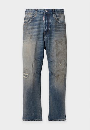 Homme debout portant un polo blanc, un jean bleu délavé, des bottes marron, une casquette marron, deux colliers croix et une ceinture avec une boucle rouge et blanche.