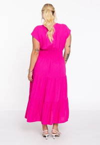 Fuchsia-maxikleid mit kurzen Ärmeln, geraffter Taille und schichtweise angeordnetem Stoff. Weiche Textur. Getragen mit weißen, schmalen Sandalen.