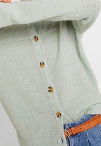 Cardigan vert menthe clair en tissu doux et texturé, avec de gros boutons marron et une coupe décontractée, associé à un jean en denim bleu.