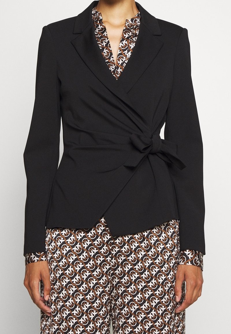 Femme portant un blazer noir à nouer à la taille sur une robe marron et blanche à motifs géométriques.
