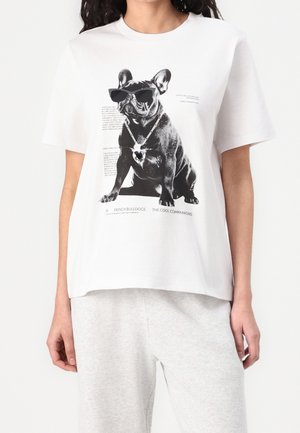 Biała koszulka z czarnym buldogiem francuskim w okularach przeciwsłonecznych i naszyjnikiem w kształcie serca, noszona przez osobę w jasno szarych spodniach.