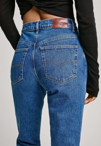 Henkilö, jolla on päällään korkeavyötäröiset siniset farkut ja musta pitkähihaisen paita, esittää selkäpuolta, jossa näkyvät taskut ja nahkainen merkkipilkku.