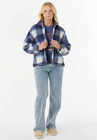 Blonde vrouw in een blauw-witte geruite jas over een marineblauwe graphic sweatshirt, lichte spijkerbroek en beige schoenen, staand met haar gezicht naar voren.