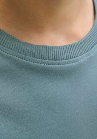 Detailní záběr na krk a límec osoby, ukazující měkkou, modro-zelenou bavlněnou košili s žebrovaným výstřihem.