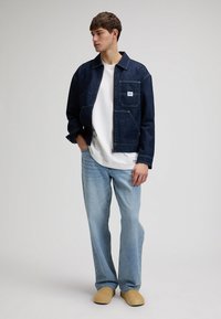 Junger Mann steht und trägt eine dunkle Jeansjacke, ein weißes T-Shirt, hellblaue Jeans und beige Slipper vor einem schlichten Hintergrund.