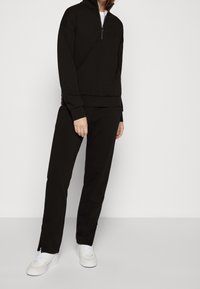 Sudadera negra con cremallera y pantalones a juego, ambos en un tejido suave. Presenta un cuello alto y puños acanalados. Con zapatillas blancas.