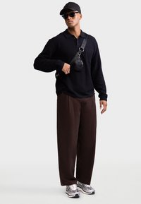 Camisola preta texturada de manga comprida, calças largas castanhas, sapatilhas prateadas, óculos de sol pretos e boné com uma bolsa transversal. Design simples e moderno.