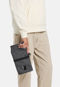 Grijze softshell tas in de hand gehouden, voorzien van een riem en een logo patch. De tas heeft een ronde vorm en een gestructureerd oppervlak.