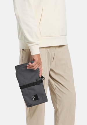 Sac à coque souple gris tenu à la main, avec une bandoulière et un patch logo. Le sac a une forme arrondie et une surface texturée.