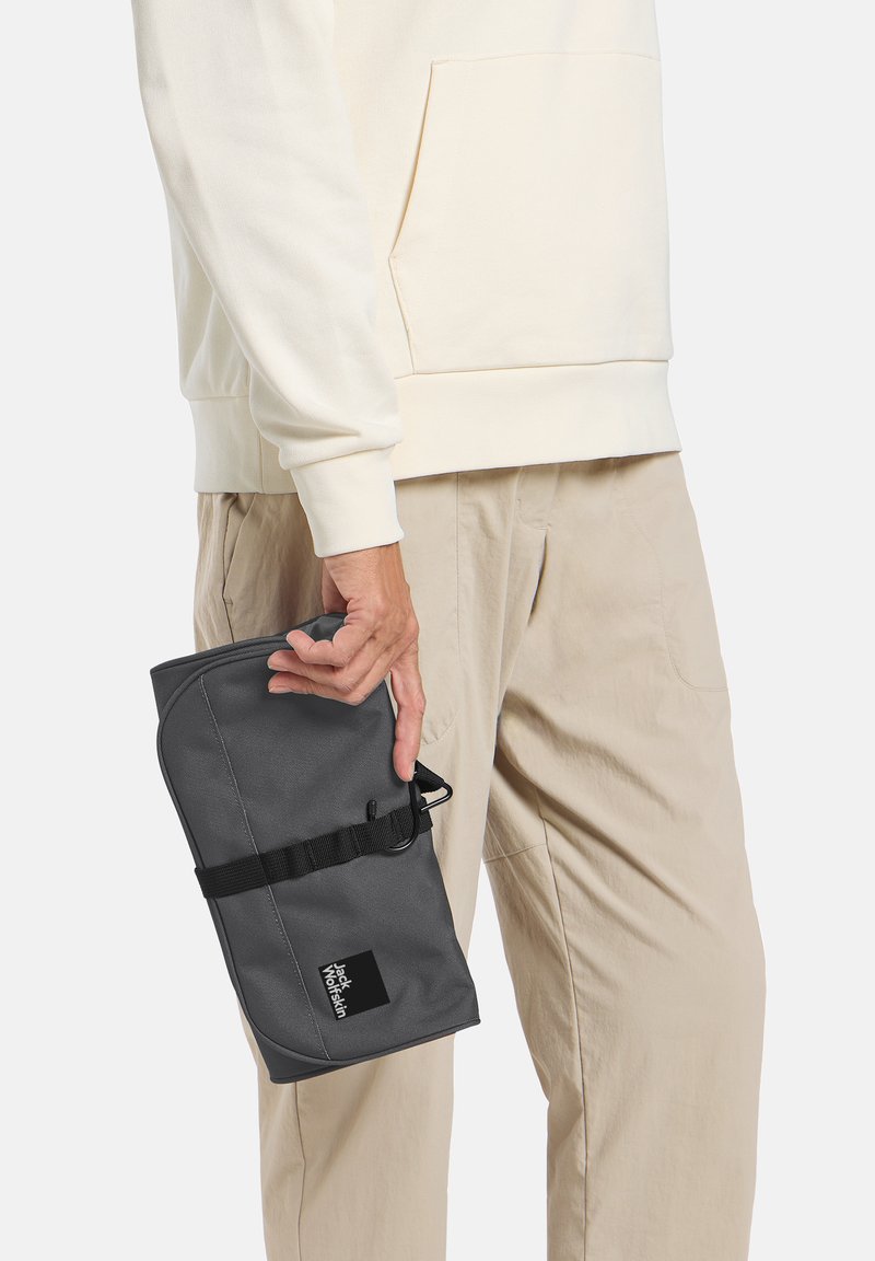 Grijze softshell tas in de hand gehouden, voorzien van een riem en een logo patch. De tas heeft een ronde vorm en een gestructureerd oppervlak.