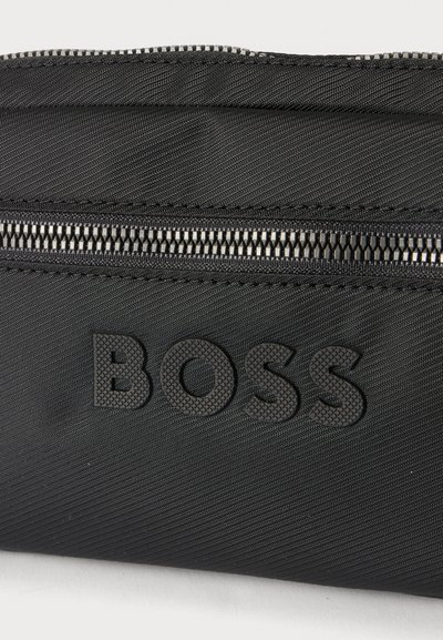 Surface en tissu noir avec des lettres majuscules texturées "BOSS" et une fermeture éclair noire partiellement ouverte au-dessus du texte.