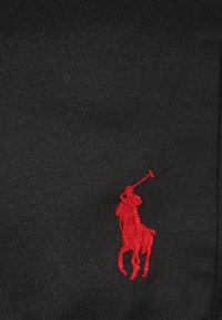 Černá tkanina s podrobným červeným vyšitým logem jezdce na polo, zdůrazňující texturu a design bez dalších ozdob.